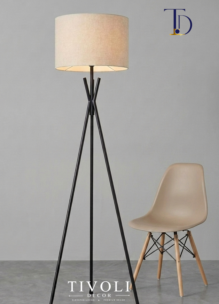 Branded Tivoli Decor minimalist metal tripod floor lamp with a matte black crossed neck frame and beige linen shade next to a modern beige shell chair.