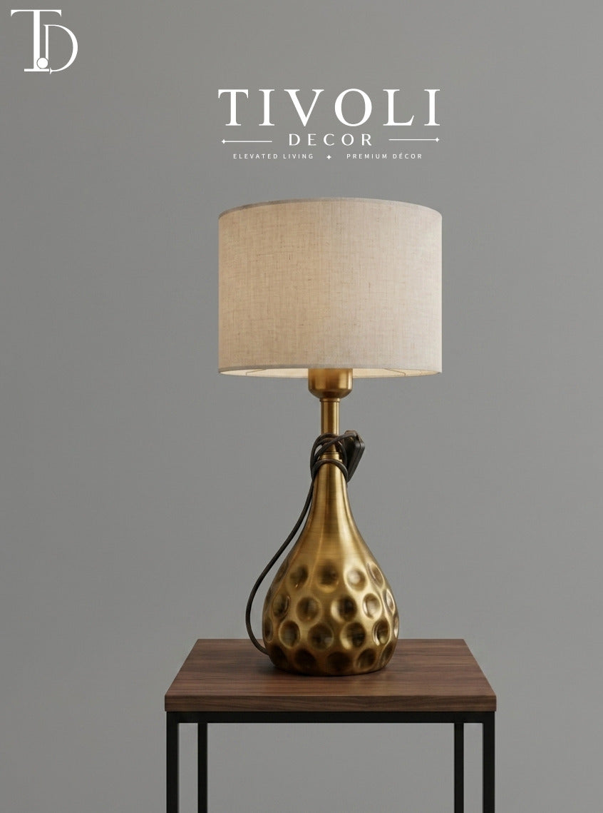 Branded Tivoli Decor luxury table lamp with a dimpled teardrop brass base and oatmeal linen shade styled on a dark wood table.