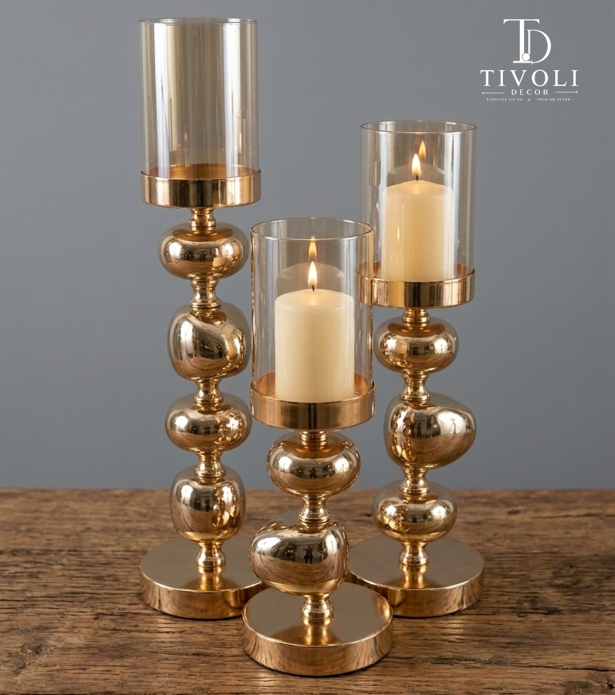 Set of 3 gold bubble-stem hurricane candle holders with lit pillar candles on wooden table – Tivoli Decor