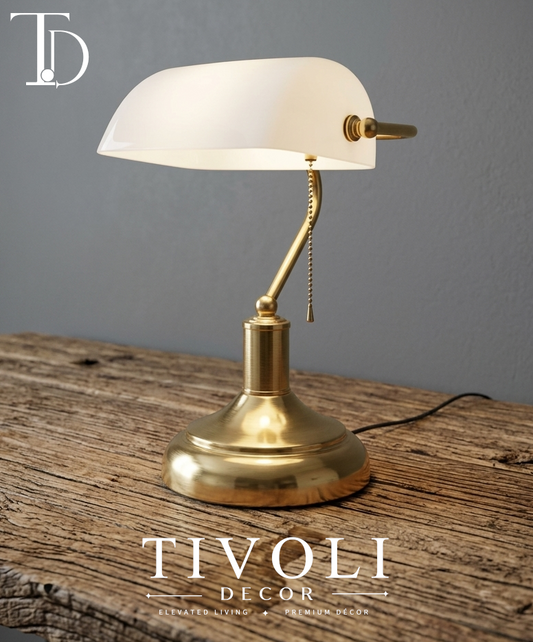 Branded Tivoli Decor luxury bankers lamp with a high gloss white glass shade and gold brushed brass base sitting illuminated on a rustic wood desk surface.