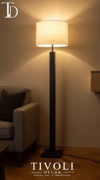 Branded Tivoli Decor modern wooden pillar floor lamp with a dark geometric column stem and cream fabric shade casting a warm glow next to a grey sofa.