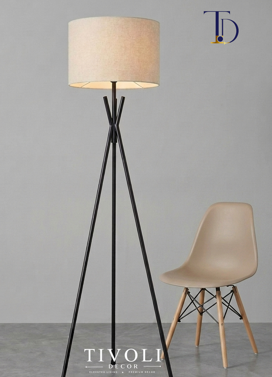 Branded Tivoli Decor minimalist metal tripod floor lamp with a matte black crossed neck frame and beige linen shade next to a modern beige shell chair.
