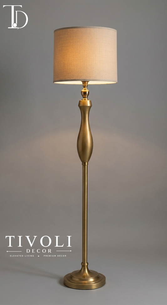 Branded Tivoli Decor luxury antique brass floor lamp with a sculpted trophy style gold stem and beige linen drum shade against a grey studio background.