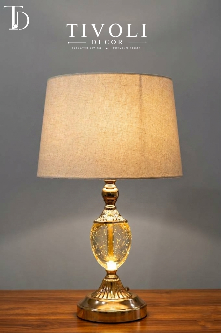 Branded Tivoli Decor luxury table lamp with an illuminated gold seeded glass bubble body, fluted gold pedestal base, and beige linen shade on a wood table.