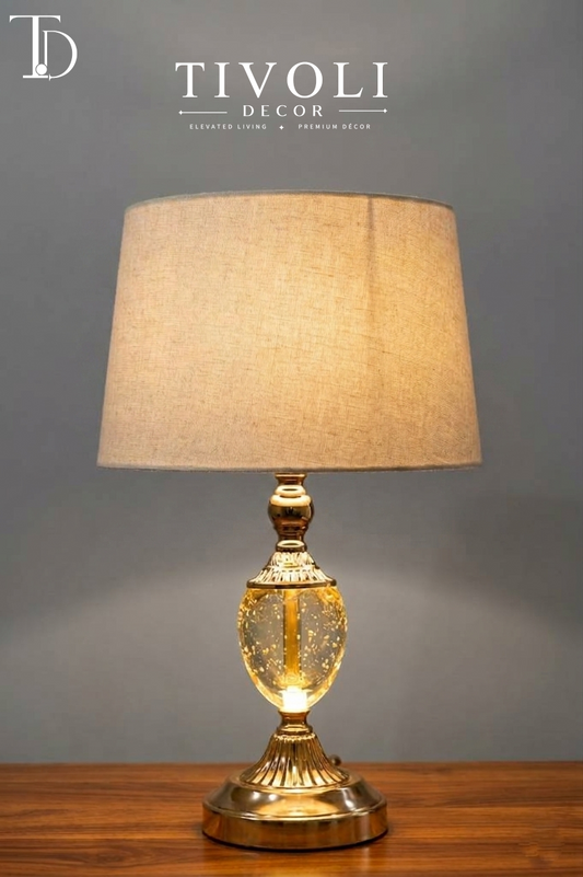 Branded Tivoli Decor luxury table lamp with an illuminated gold seeded glass bubble body, fluted gold pedestal base, and beige linen shade on a wood table.